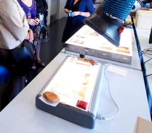 Photographie en couleur de tables lumineuses sur une table. Des négatifs de nitrate sont disposés sur les tables lumineuses. À l’arrière-plan : trois personnes se tiennent près de la table.