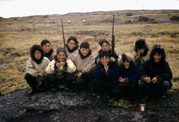 Photo couleur d’un groupe de garçons inuits accroupis sur un rocher plat; deux d’entre eux ont un fusil dans les mains