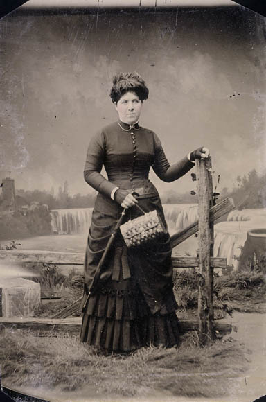 Ferrotype noir et blanc d’une femme posant debout devant une clôture de bois avec derrière elle une toile de fond représentant les chutes du Niagara.