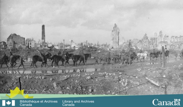 Photo noir et blanc d’une ville ravagée par la guerre. Une colonne de troupes composée majoritairement d’hommes à cheval traverse la ville.