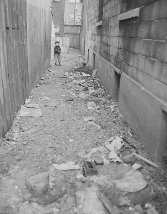Photographie en noir et blanc montrant une ruelle longue et étroite jonchée de déchets. Un jeune enfant se tient à l’autre extrémité de la ruelle et regarde le photographe.