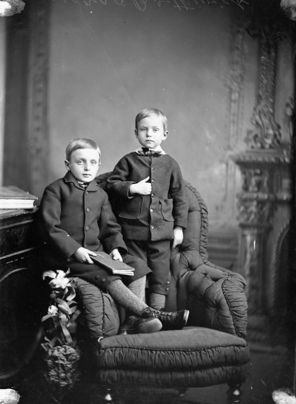 Photographie en noir et blanc de deux jeunes garçons portant des vestes noires, l'un est assis sur l'accoudoir d'une chaise et l'autre est debout sur la même chaise.