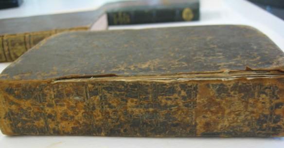 Photographie en couleur montrant plusieurs livres sur une table. Le livre à l'avant-plan présente un cuir extrêmement détérioré, et le dos du livre s'est détaché de la couverture.