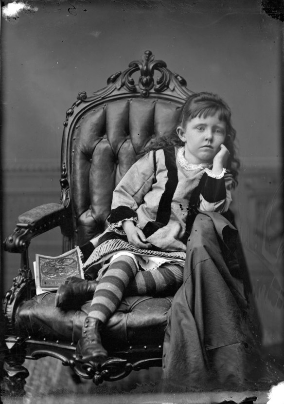 Photographie en noir et blanc d'une petite fille, le menton appuyé sur la main, un livre à côté d'elle.