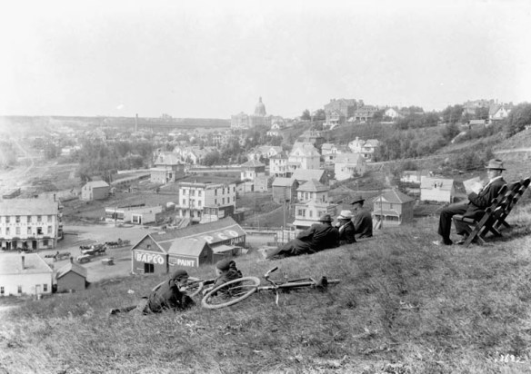 Une photographie en noir et blanc prise à partir d’une colline surplombant une ville; on y voit des cyclistes qui se reposent couchés dans l’herbe et, à proximité, d’autres hommes sont assis.