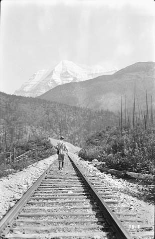 Une photographie en noir et blanc montrant un homme marchant sur la voie ferrée, en direction du mont Robson, qu’on distingue au loin.  