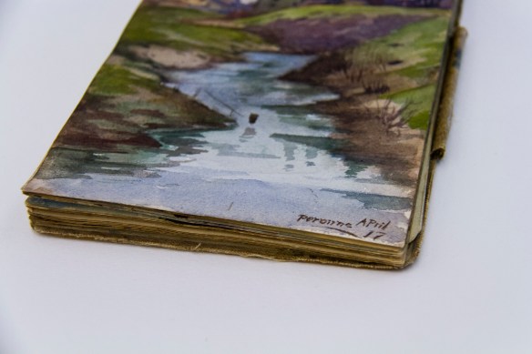 Une photographie couleur d’un cahier de dessins vu de biais, montrant les berges d’une rivière avec la date et le lieu dans le coin inférieur droit, « Perrone avril 17 ».