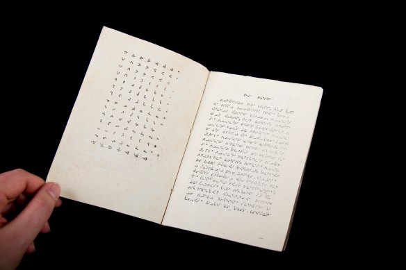 Image en couleur d’un livre écrit en caractères syllabiques inuktitut. Le livre est ouvert pour montrer les deux premières pages. Celle de gauche présente le syllabaire inuktitut et celle de droite présente un texte rédigé en syllabique inuktitut. 