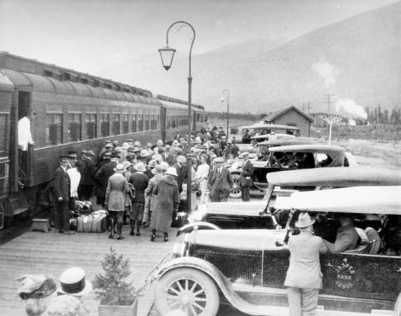 Photographie en noir et blanc d’une foule et de bagages sur le quai, à côté d’un train de passagers. Deux porteurs se trouvent à côté du train. Le premier est sur le quai et veille sur les bagages tandis que l’autre est debout à la porte de la voiture. Au premier plan se trouve une automobile portant la mention « Jasper Park Lodge » sur la portière. Des montagnes sont visibles au loin.