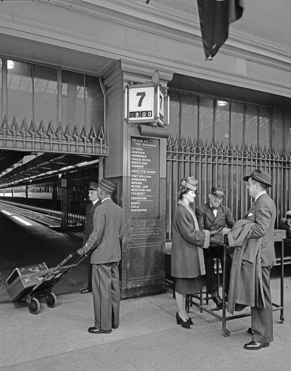 Photographie en noir et blanc de personnes à une gare. Un porteur transportant des bagages sur un chariot est vu de dos. Deux passagers élégamment vêtus discutent avec un préposé aux billets. Sur le mur derrière eux, un panneau d’information annonce le train Dominion, qui part de Montréal pour se rendre à Vancouver. Un train de voyageurs est visible à l’arrière-plan.