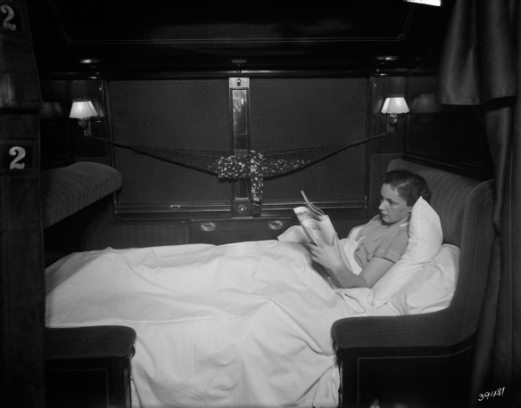 Photographie en noir et blanc d’une femme de profil, étendue sous les couvertures de la couchette inférieure, lisant un journal.