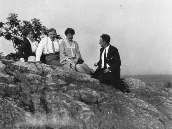Photo en noir et blanc d’un groupe de personnes assises sur une haute barre rocheuse, devant des arbres flous et un arrière-plan distant. Chacun regarde en direction du photographe, sauf un homme qui regarde les autres.