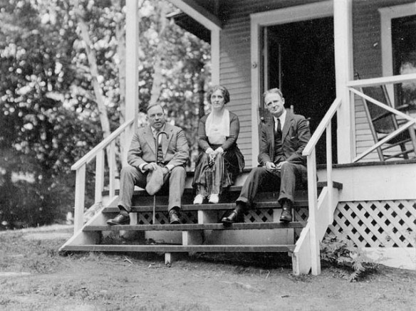 Photo en noir et blanc de trois personnes assises sur les marches d’escalier d’un chalet et regardant en direction du photographe. 