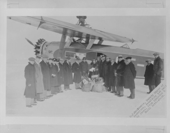 Une photographie en noir et blanc illustrant un groupe d’hommes vêtus de manteaux d’hiver et de chapeaux devant un avion monomoteur. Les hommes sont debout, en demi-cercle, et font face à l’appareil-photo. Des sacs de courrier-avion reposent sur le sol.