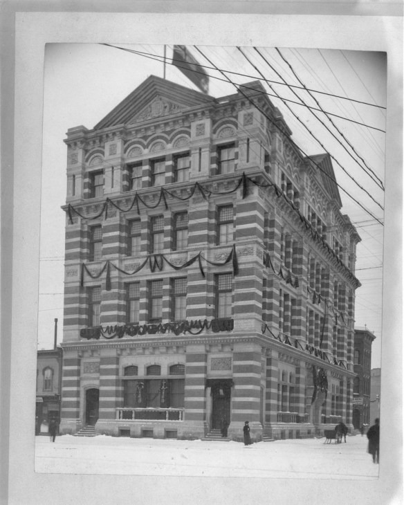 Photo en noir et blanc d’un édifice décoré pour Noël, bordé d’une rue enneigée. La façade, très travaillée, révèle plusieurs couches alternées de pierres et de briques.