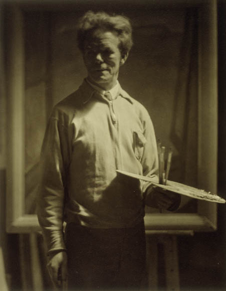 Photographie en noir et blanc montrant un homme, debout, tenant une de la peinture et des pinceaux dans une main avec le regard un peu à l’écart.