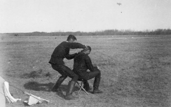 Photographie en noir et blanc d’un soldat qui coupe les cheveux d’un soldat assis dans un champ.