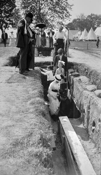 Photo noir et blanc montrant des hommes, des femmes et des enfants dans de beaux habits qui explorent et observent une tranchée canadienne reconstituée dans le cadre d’une exposition en plein air.