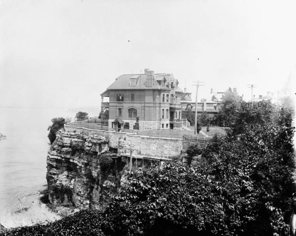 Photographie en noir et blanc d’une grande maison de brique perchée au sommet d’une falaise.