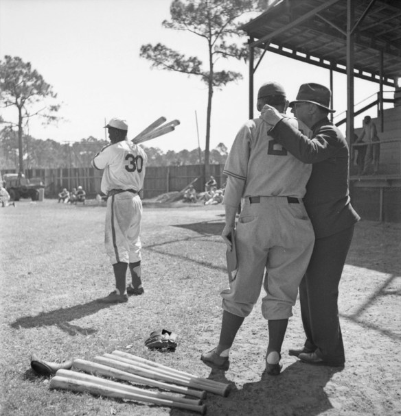 Photographie en noir et blanc de trois hommes sur un terrain de baseball. Deux hommes, dont l'un est vêtu d'un uniforme et l'autre porte une tenue de ville, sont debout en avant-plan et se parlent. Sur le sol, à leurs pieds, on peut voir deux gants de baseball et des bâtons de baseball. En arrière-plan, un autre joueur, en uniforme, tient trois bâtons de baseball reposant sur son épaule droite. Au loin, des gens sont debout ou assis sur le sol ou sur les bancs-gradins.