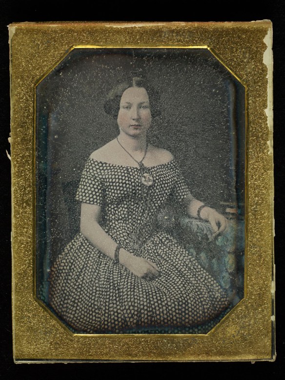 Photographie en couleur dans un cadre doré montrant une femme assise avec le bras accoudé sur une petite table de chevet. Le verre est à un stade avancé de détérioration. On voit le phénomène de transpiration du verre; l’image est floue.