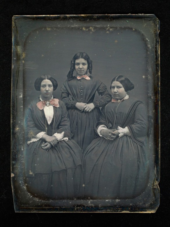 Daguerréotype coloré à la main. Image de trois jeunes femmes; celle du milieu est debout entre les deux autres, assises.