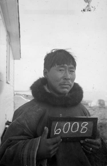 Une photographie en noir et blanc d’un homme inuit tenant un petit tableau sur lequel est écrit le numéro 6008.