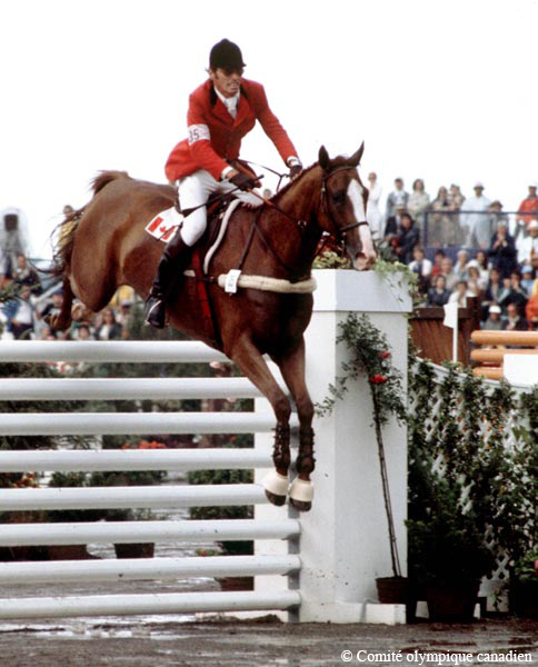 Photographie en couleur d’un homme à cheval qui franchit un obstacle durant une compétition équestre. En arrière-plan, des spectateurs sont assis.