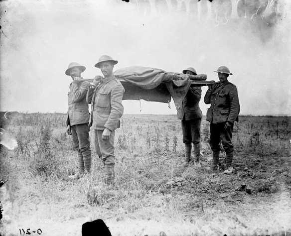 Photographie en noir et blanc de quatre soldats transportant une civière sur laquelle repose un corps recouvert d’une bâche, dans un paysage de désolation.
