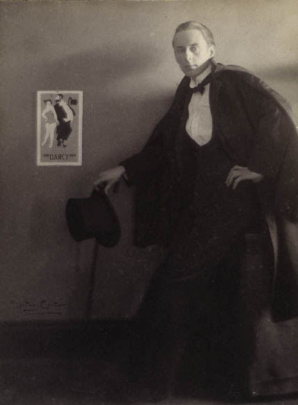 Photo noir et blanc d’un homme en tenue de soirée, debout à côté d’une affiche posée sur un mur.