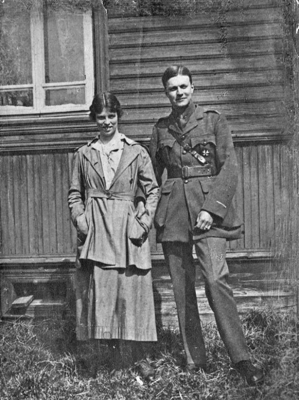 Une photographie en noir et blanc montrant un couple debout devant un bâtiment en bois. Elle porte ce qui semble être un uniforme d’infirmière et lui, un uniforme de l’armée russe.