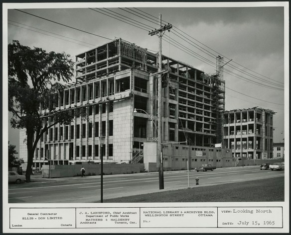 Photographie en noir et blanc de la façade d’un édifice de dix étages en construction. On voit des panneaux de construction à la hauteur du rez-de-chaussée, des automobiles (y compris une Coccinelle de Volkswagen!) et des piétons.