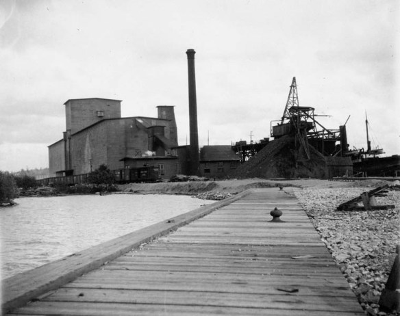 Une photographie en noir et blanc montrant un long quai en bois près d’un rivage et des silos à grains à l’arrière-plan
