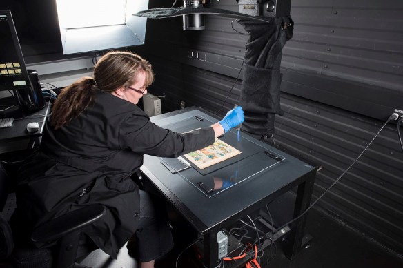 Une bande dessinée est placée sur une surface plane noire sous une feuille de plexiglas. Une femme est penchée sur la surface; elle enlève la poussière du plexiglas à l’aide d’un souffleur antistatique. La lentille d’un appareil photo est visible au-dessus de la table. 
