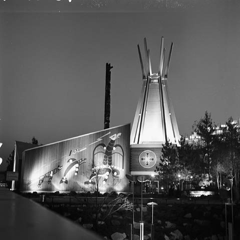 Une photographie en noir et blanc d’un édifice dont la façade est décorée de motifs d’art autochtone, contigu à une structure en forme de tipi. On aperçoit le haut d’un mât totémique derrière l’édifice.