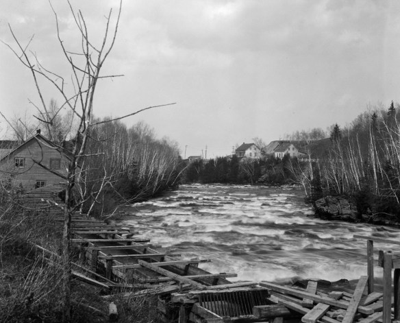  Une photographie en noir et blanc montrant une rivière avec un fort courant, des arbres de chaque côté, et un vieux moulin au fond à gauche