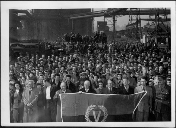 Photographie noir et blanc montrant un regroupement de personnes tenant un drapeau orné d’un « V » signifiant Victoire.