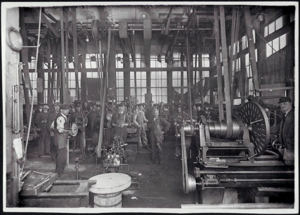 Photographie noir et blanc montrant des hommes dans une usine. On peut y apercevoir en arrière-plan un nombre important de travailleurs faisant fonctionner manuellement les premières machines mécaniques.