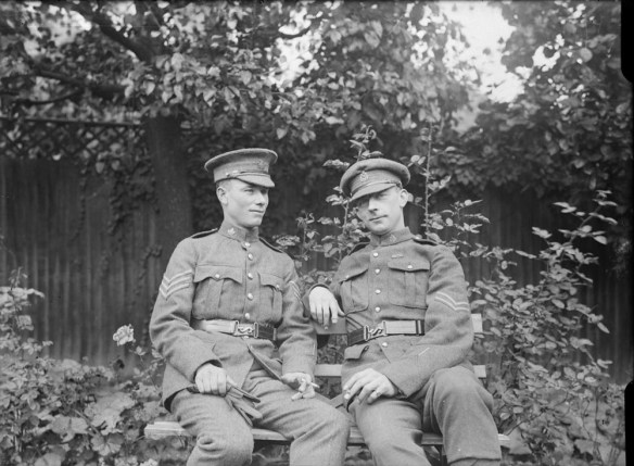 Photographie en noir et blanc de deux soldats en uniforme assis sur un banc. L’homme à droite regarde directement la caméra et arbore un sourire discret.