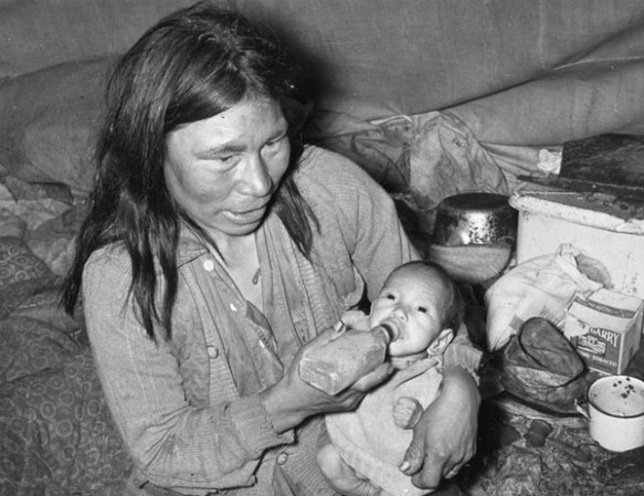 Photo noir et blanc d’une femme inuite amaigrie assise dans une petite tente avec son bébé, devant des provisions. La femme nourrit son bébé à l’aide d’un biberon rectangulaire.