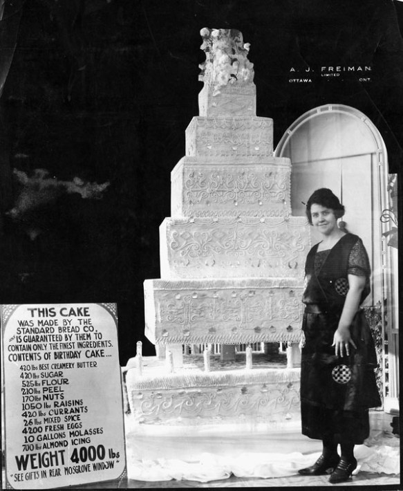 Photo noir et blanc d’un énorme gâteau avec une jeune femme debout à sa gauche. De l’autre côté, une affiche montre la liste des ingrédients.