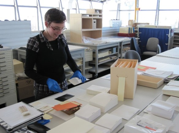 Photo couleur d’une femme portant des gants de nitrile qui retire un négatif d’une enveloppe. La table devant elle est couverte d’enveloppes et de boîtes d’archives.