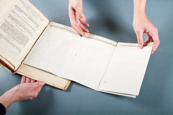 Une photo en couleur d’un livre ouvert avec une carte plié que deuux mains commencent à déplier.