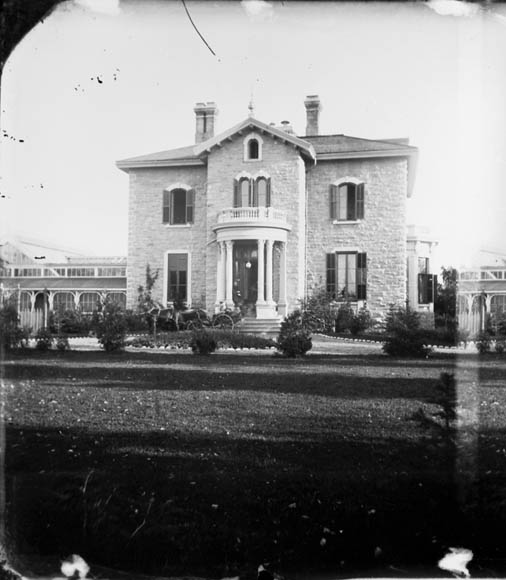 Photographie noir et blanc d’une vaste et majestueuse demeure, dont la porte centrale est flanquée de larges fenêtres. Une calèche se trouve dans l’entrée.