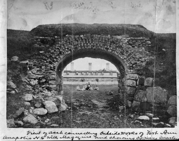 Photographie en noir et blanc d’une arche en pierre et vue par celle-ci, en arrière-plan, d’un grand immeuble clôturé avec terrain à l’avant. Une femme et cinq enfants apparaissent au centre, assis sur le sol.