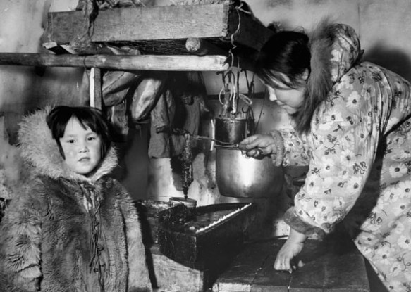 Photo noir et blanc d’une femme inuite dans un igloo qui porte une parka à motif floral et manipule une lampe à l’huile de phoque. Un jeune inuit portant une parka en fourrure se trouve près d’elle. 