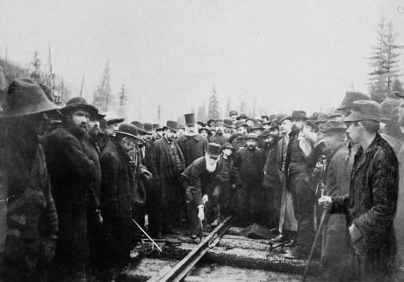 Photographie noir et blanc d’un homme martelant un rail de chemin de fer. Il est entouré d’un groupe d’hommes, dont certains fixent l’appareil photo et d’autres regardent les rails.