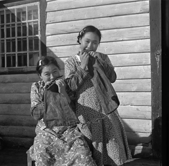 Photo noir et blanc de deux filles inuites, une debout et l’autre assise, devant une maison en bois. Elles mâchent des pièces de peau de phoque en forme de bottes.