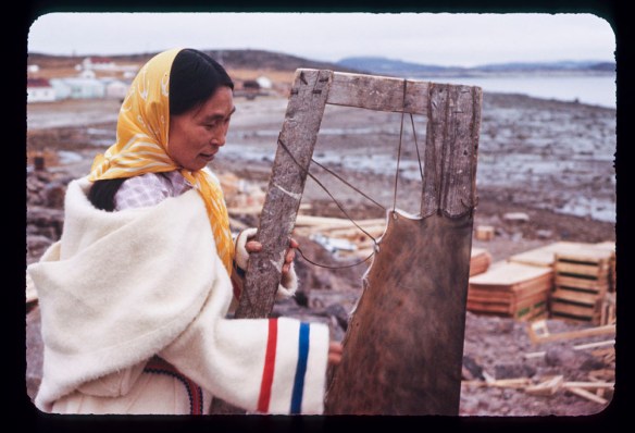 Photo en couleurs d’une femme inuite portant une parka en laine blanche et un foulard jaune. Elle se trouve près du littoral et étend une peau de phoque sur un cadre en bois. 