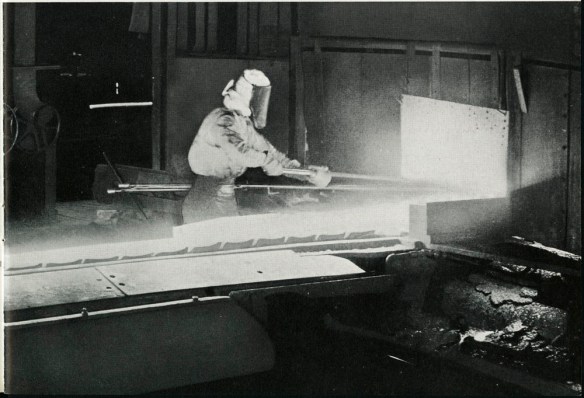 Photographie noir et blanc montrant un ouvrier manipulant une barre de fer chauffée.